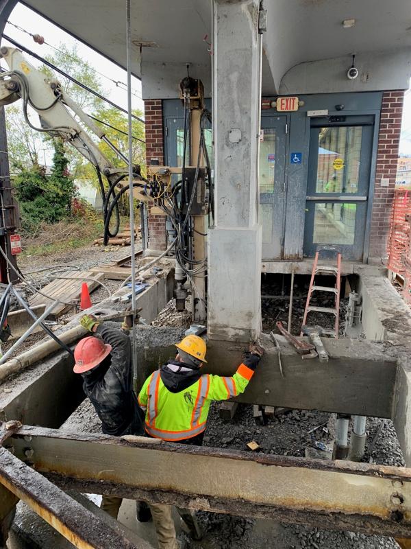 Construction workers reinforcing a concrete column under the W. 117th Street Bridge using heavy equipment and tools.