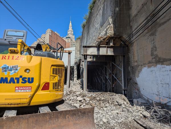 A wide shot of a construction site featuring a yellow excavator, rubble, and exposed tunnel structure. Office buildings and a tower are visible in the background.